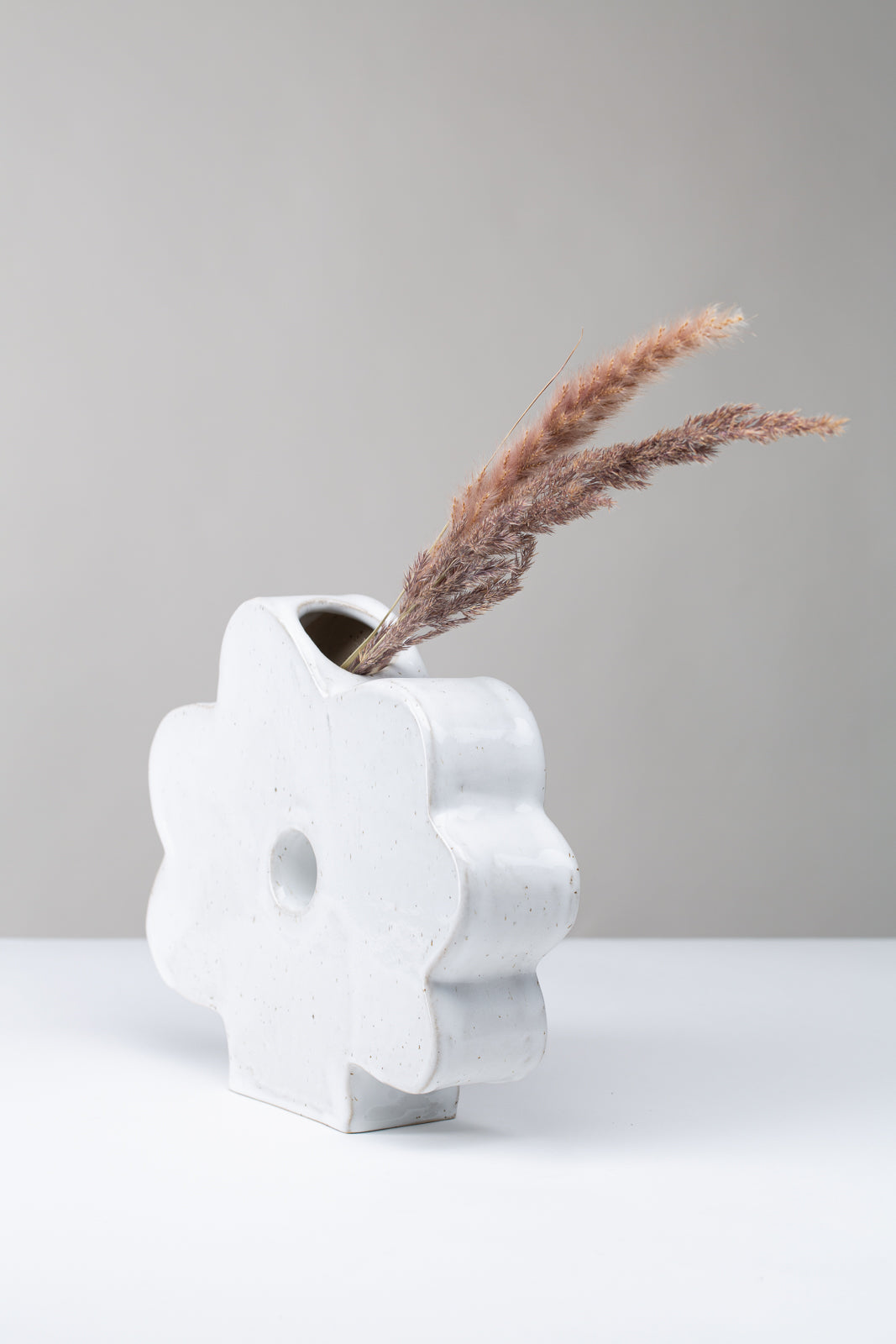 Man sieht eine Vase in Form einer Blume auf weißem Hintergrund, die Vase ist weiß mit Pünktchen, in der Vase stecken seitlich getrocknete Gräser, man sieht die Vase von der Seite