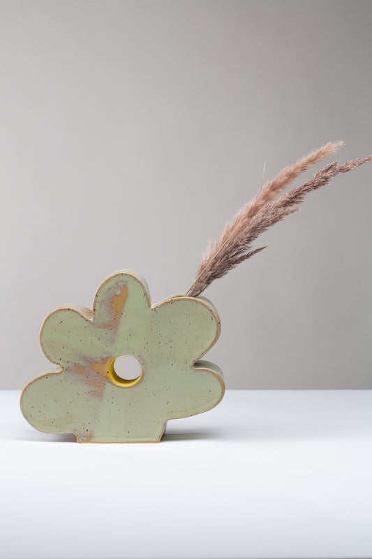 Man sieht eine Vase in Form einer Blume auf weißem Hintergrund, die Vase ist hellgrün mit Pünktchen und einem gelben Akzent ind er Mitte, in der Vase stecken seitlich getrocknete Gräser 