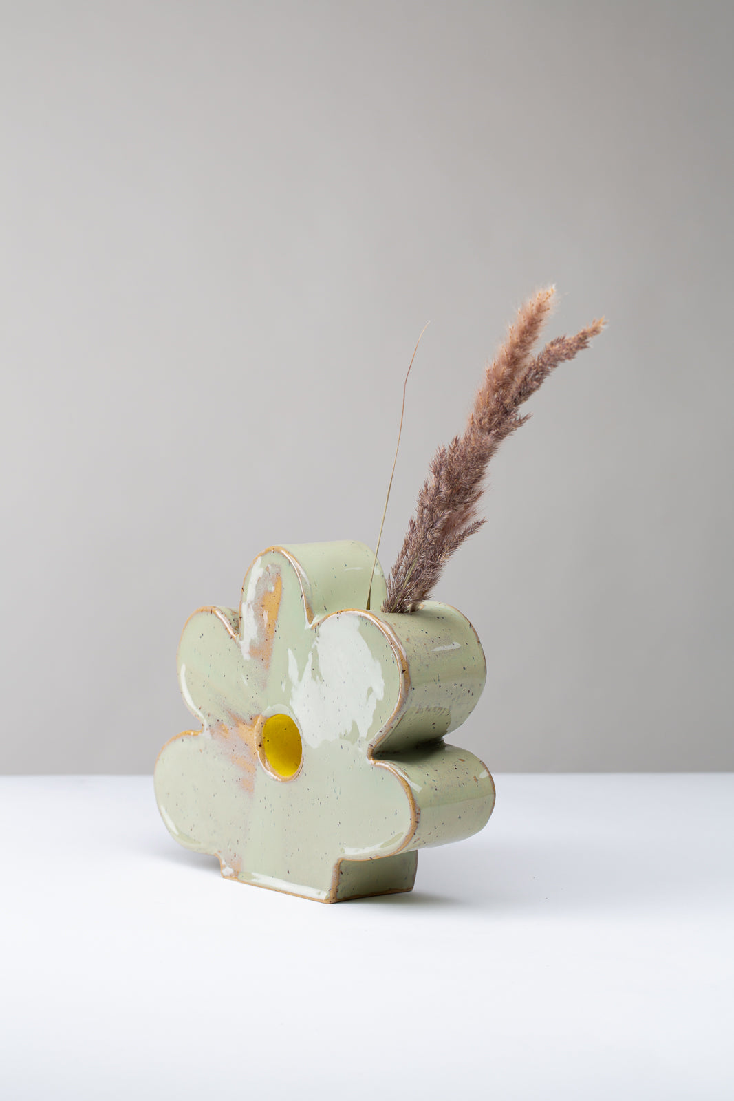 Man sieht eine Vase in Form einer Blume auf weißem Hintergrund, die Vase ist hellgrün mit Pünktchen und einem gelben Akzent ind er Mitte, in der Vase stecken seitlich getrocknete Gräser, man sieht die Vase von der Seite